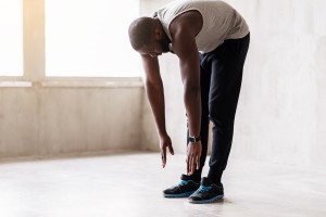 Concentrated bearded sportsman is standing on straight legs and making forward bend. Focus on smart watch and sport shoes